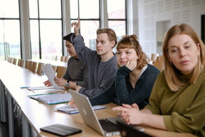 Studierende im Seminarraum sitzen an den Tischen. Eine Person meldet sich. 